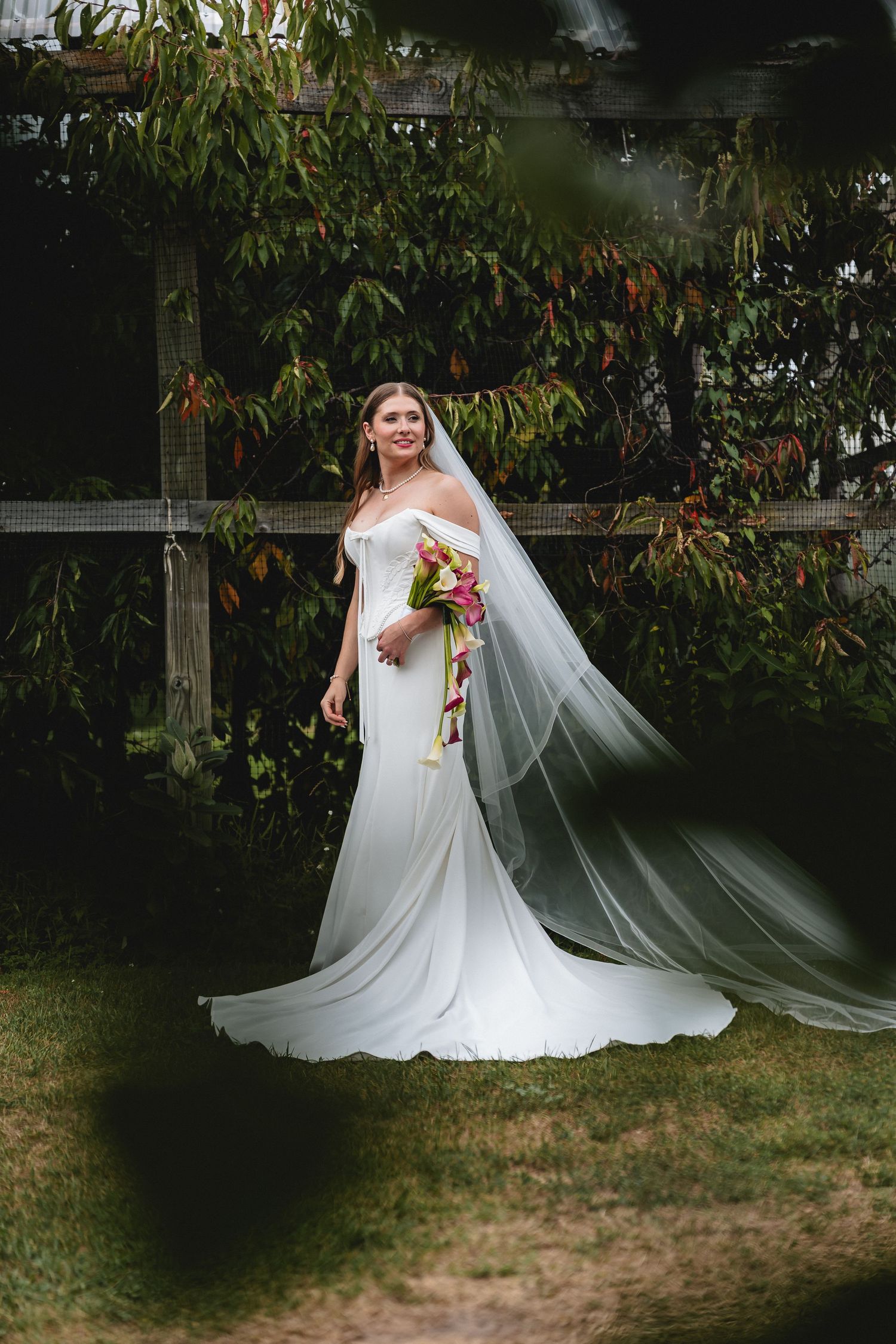 Bride in white off-shoulder gown with flowing veil holds colorful bouquet in lush garden setting.