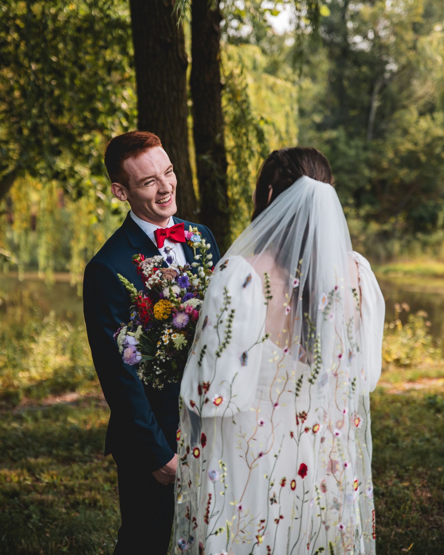 Groom in navy suit with red bow tie laughs at bride in floral embroidered wedding dress and veil outdoors.