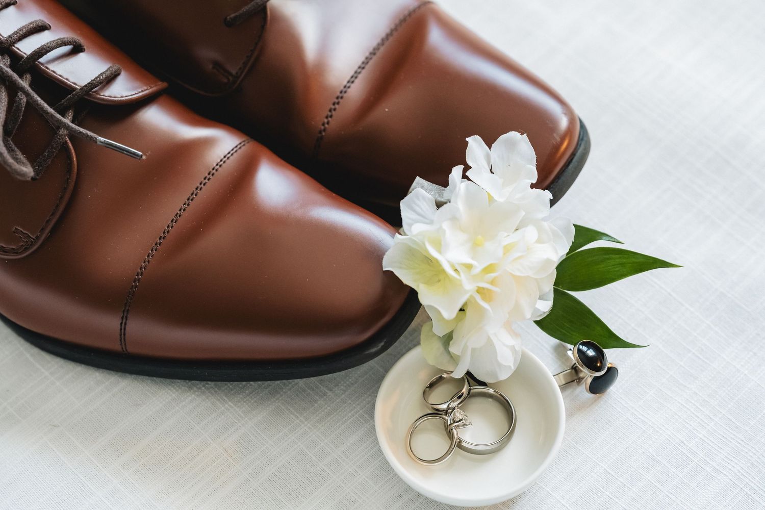 Brown leather dress shoes with white floral boutonniere and wedding rings on a white surface.