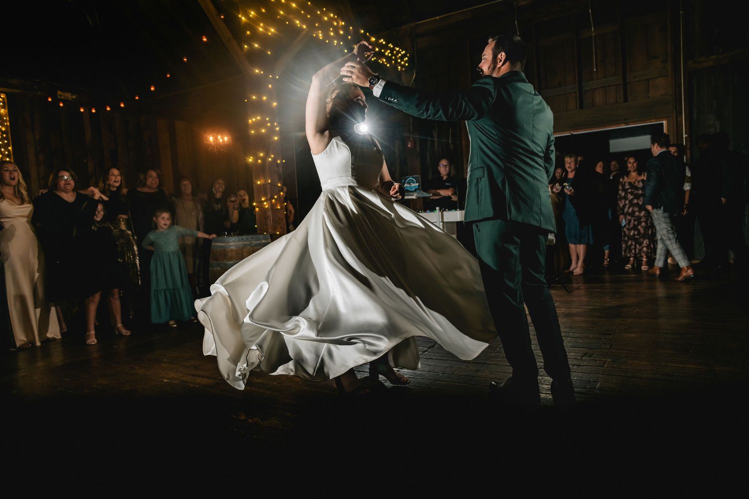 Bride and groom share first dance at wedding reception, with flowing white gown and warm string lights in background.