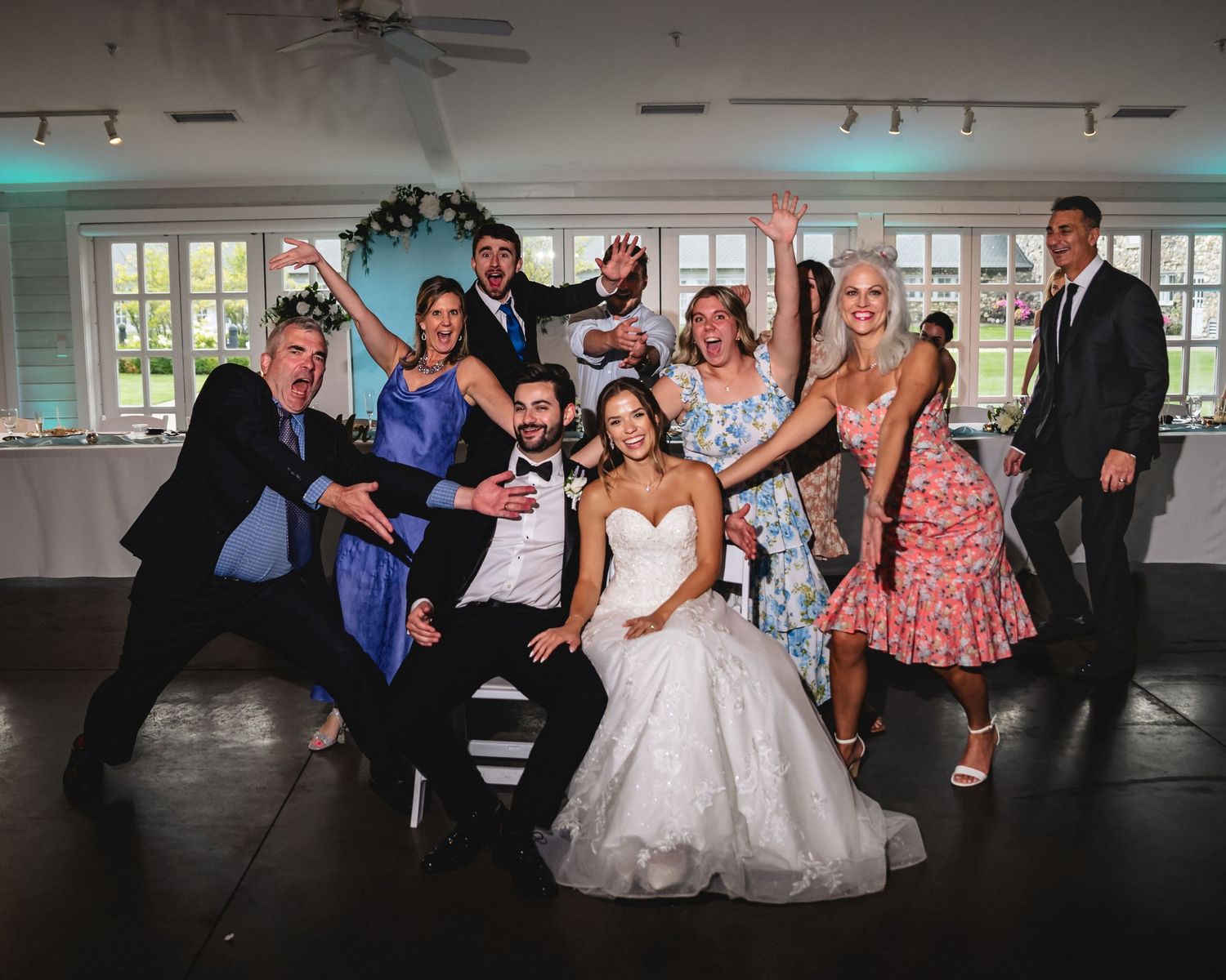 Bride and groom pose with joyful wedding guests on dance floor at elegant indoor reception venue.
