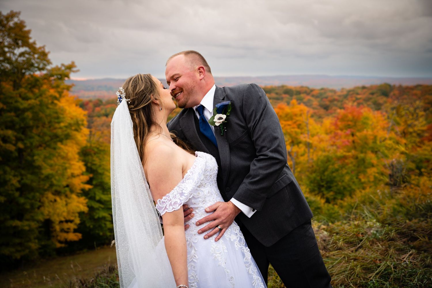 A couple shares a romantic kiss on a hilltop overlooking vibrant autumn foliage on their wedding day.