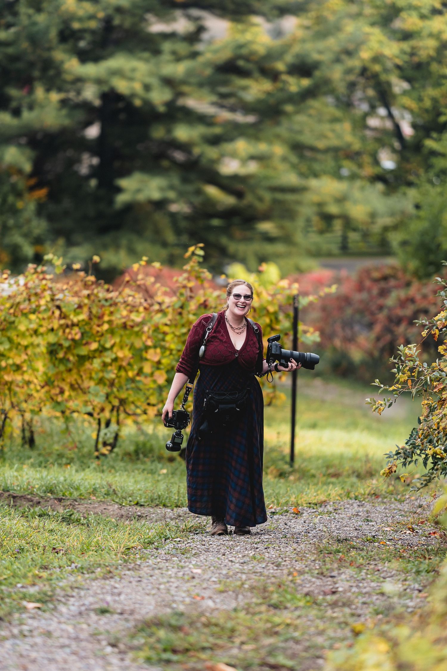 A person in dark clothing walks along a gravel path bordered by autumn foliage and trees in a garden setting.