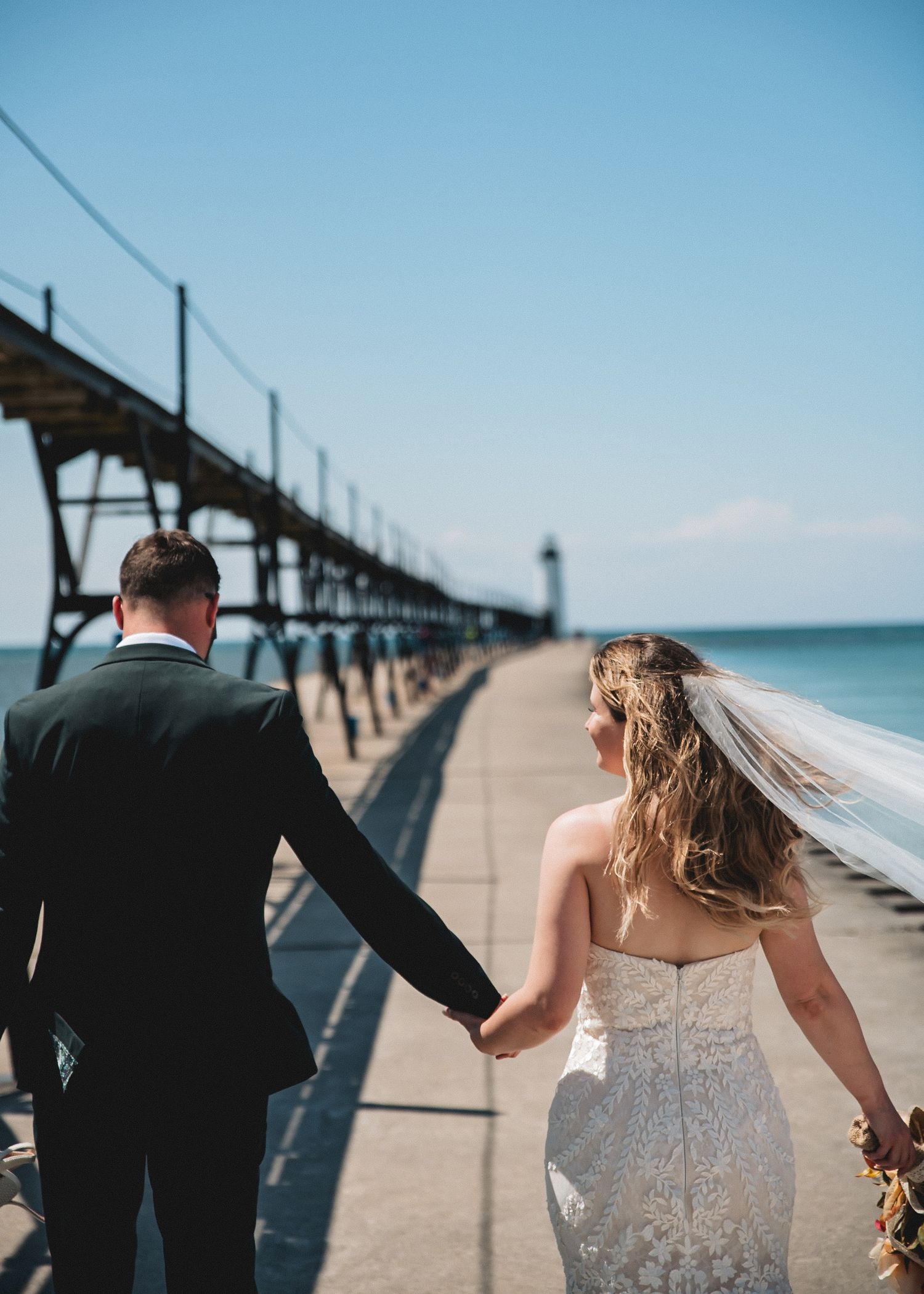 A bride and groom walk hand-in-hand along a pier near the lighthouse in Manistee MI on a sunny wedding day.