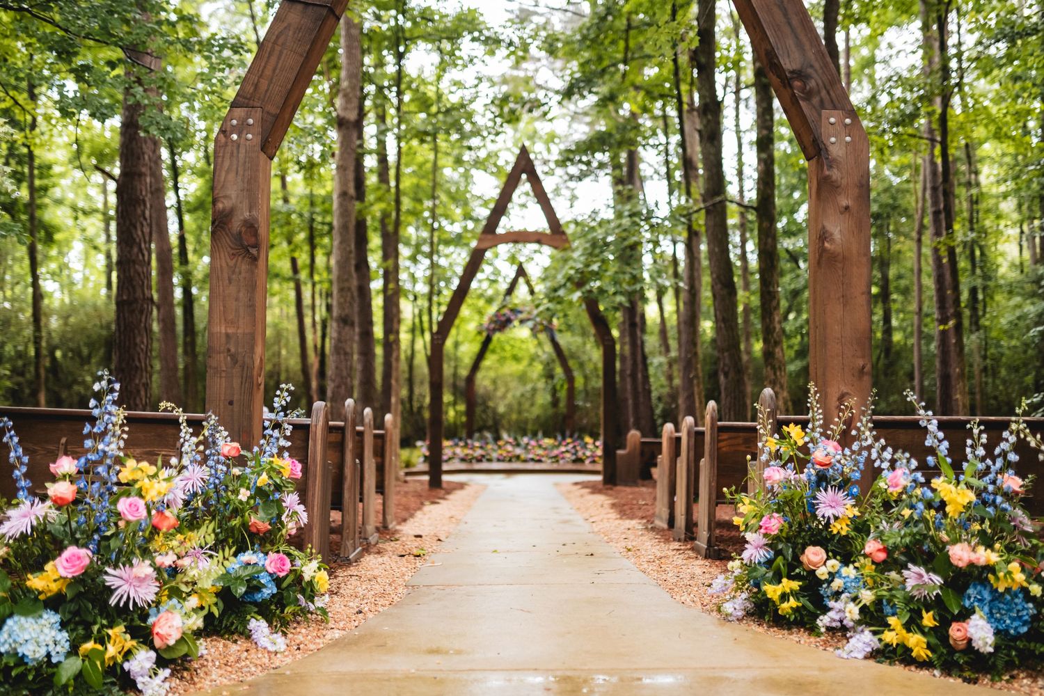 A tree-lined pathway decorated with colorful floral arrangements creates a romantic outdoor wedding ceremony setting.