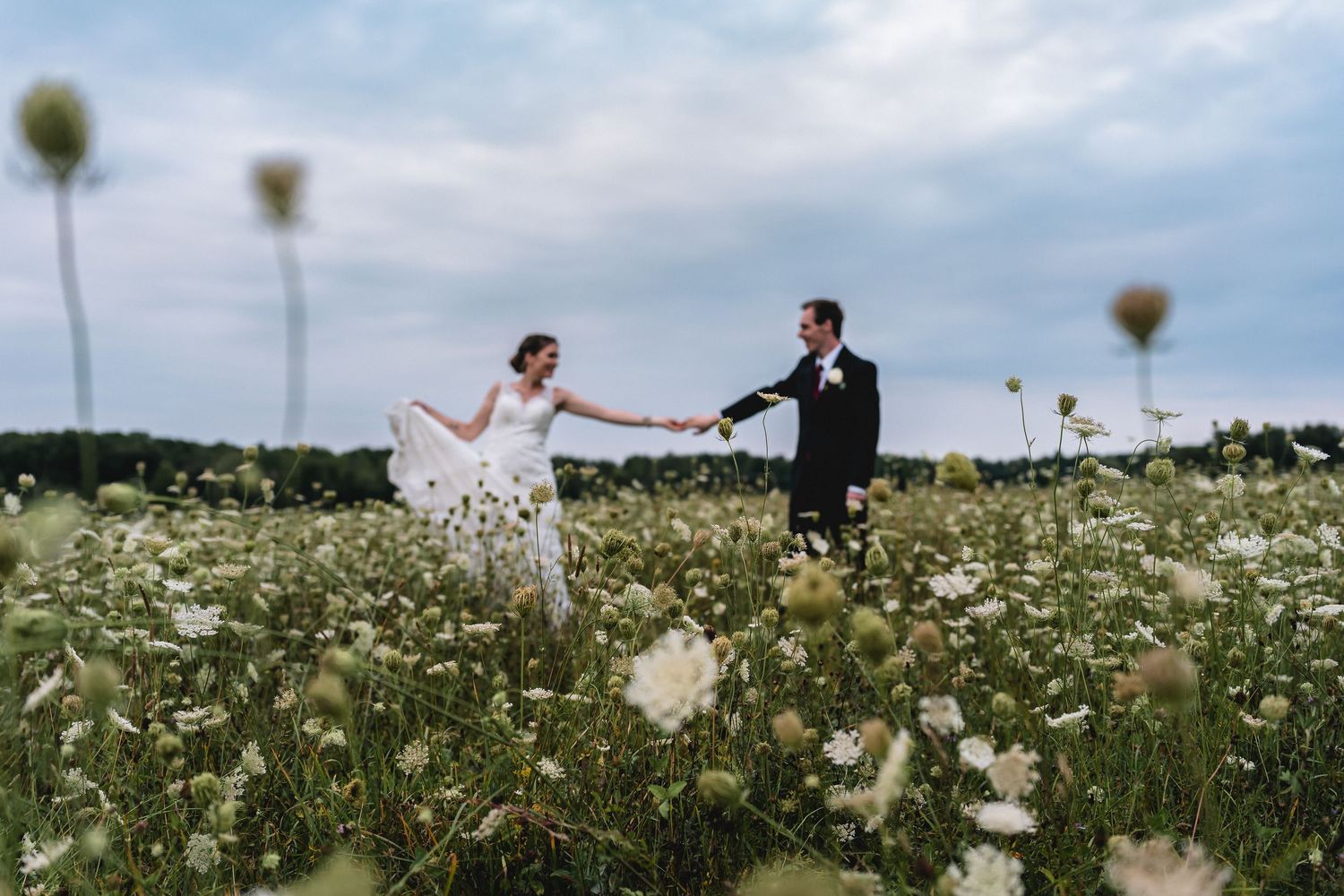 Bride and groom holding hands in a wildflower meadow on their wedding day under a cloudy sky.