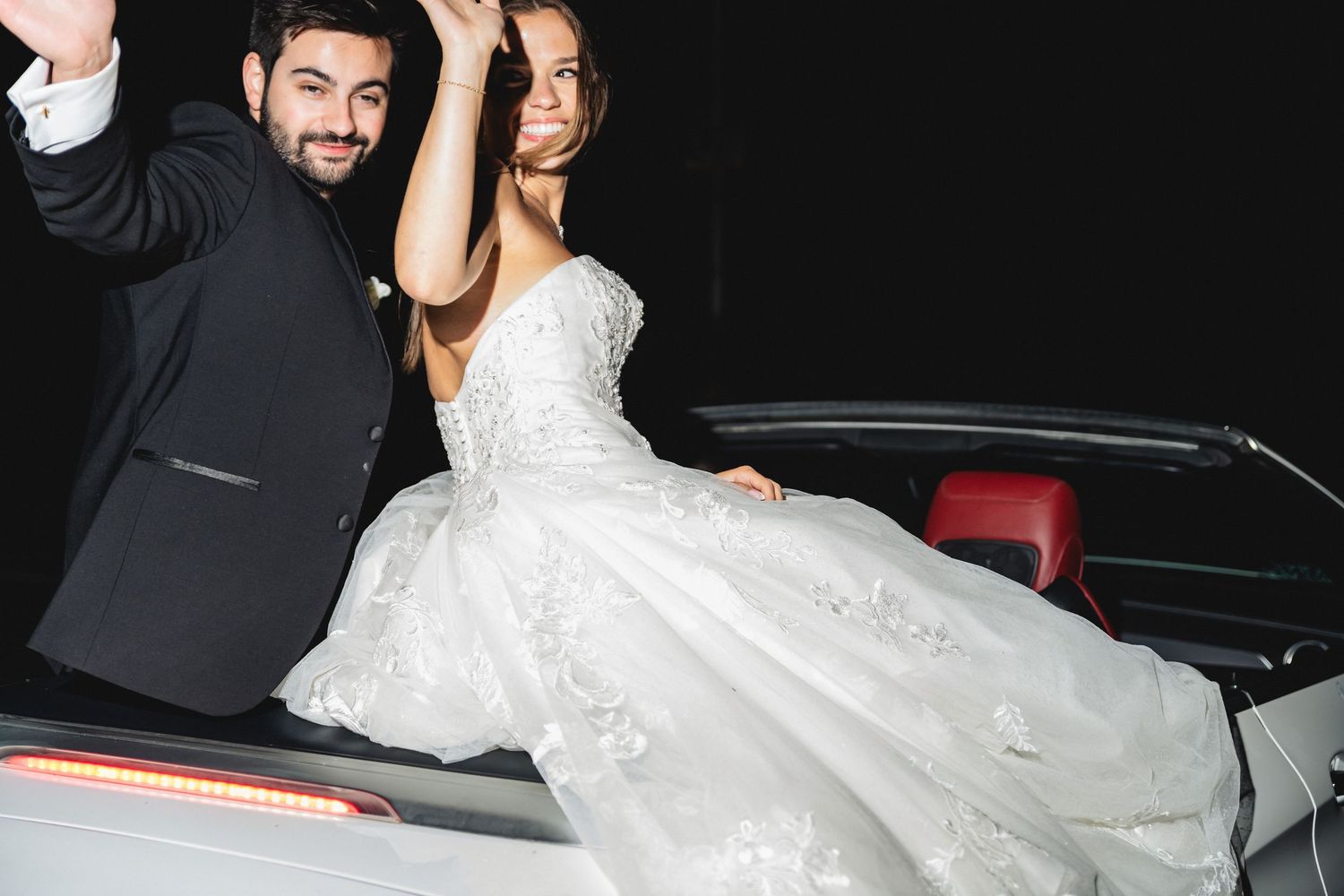 Wedding party poses next to a luxury vehicle during evening celebrations.