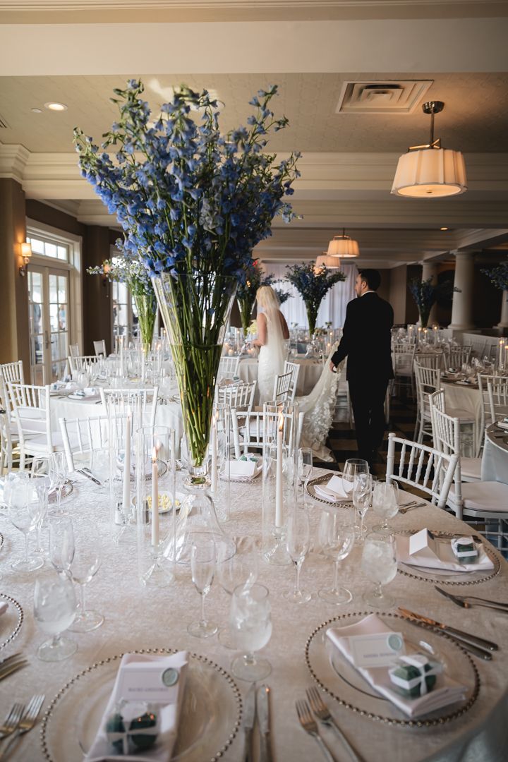 Elegant wedding reception tables set with white linens, crystal glassware and tall blue delphinium centerpieces.