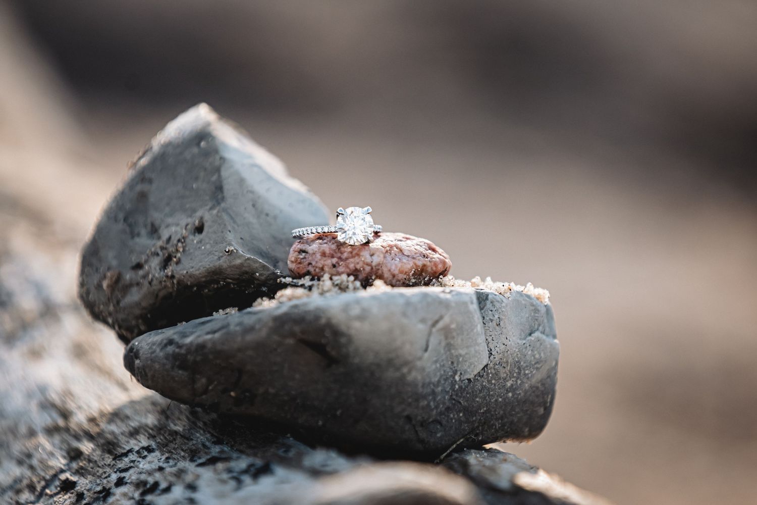 A diamond engagement ring resting on weathered gray rocks outdoors.