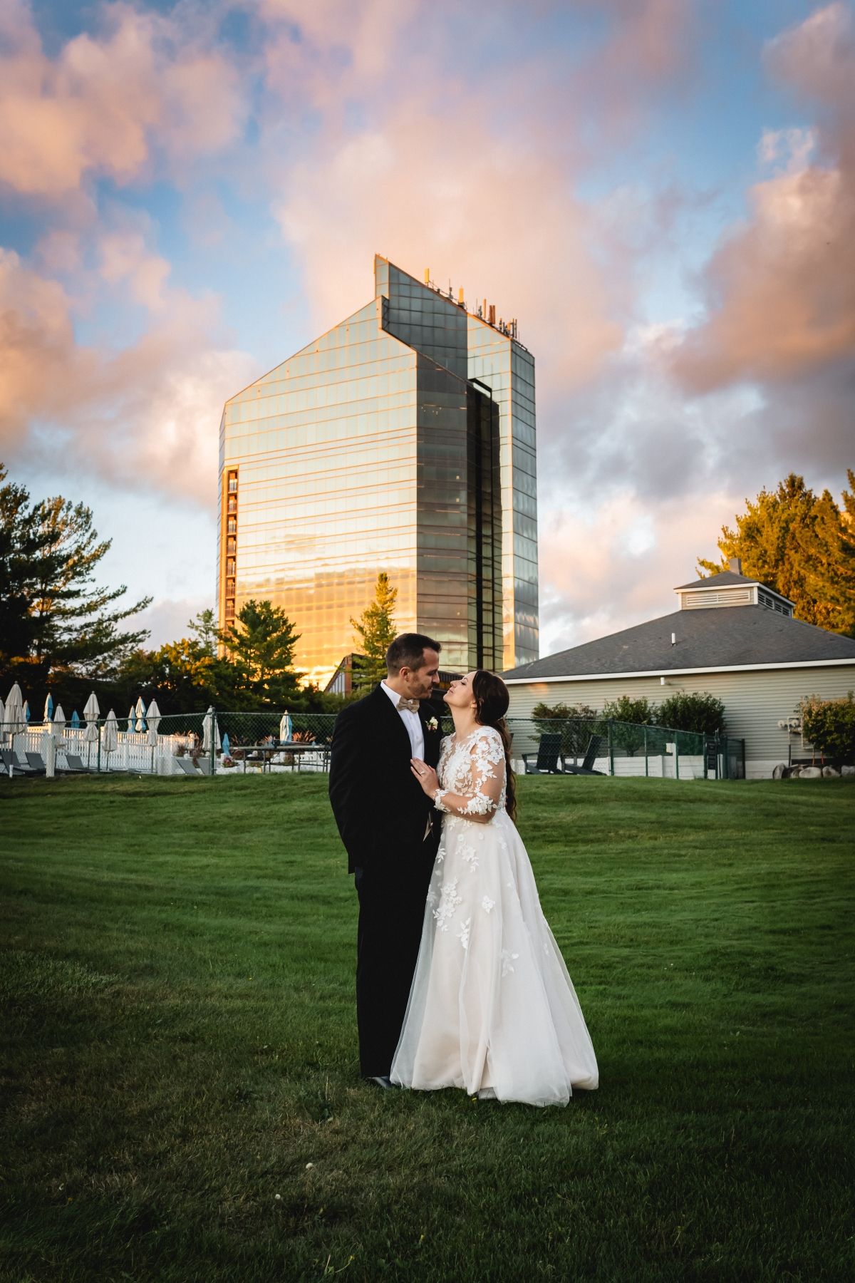 A couple in wedding attire stands on a grassy lawn at sunset with a modern glass office building in the background.
