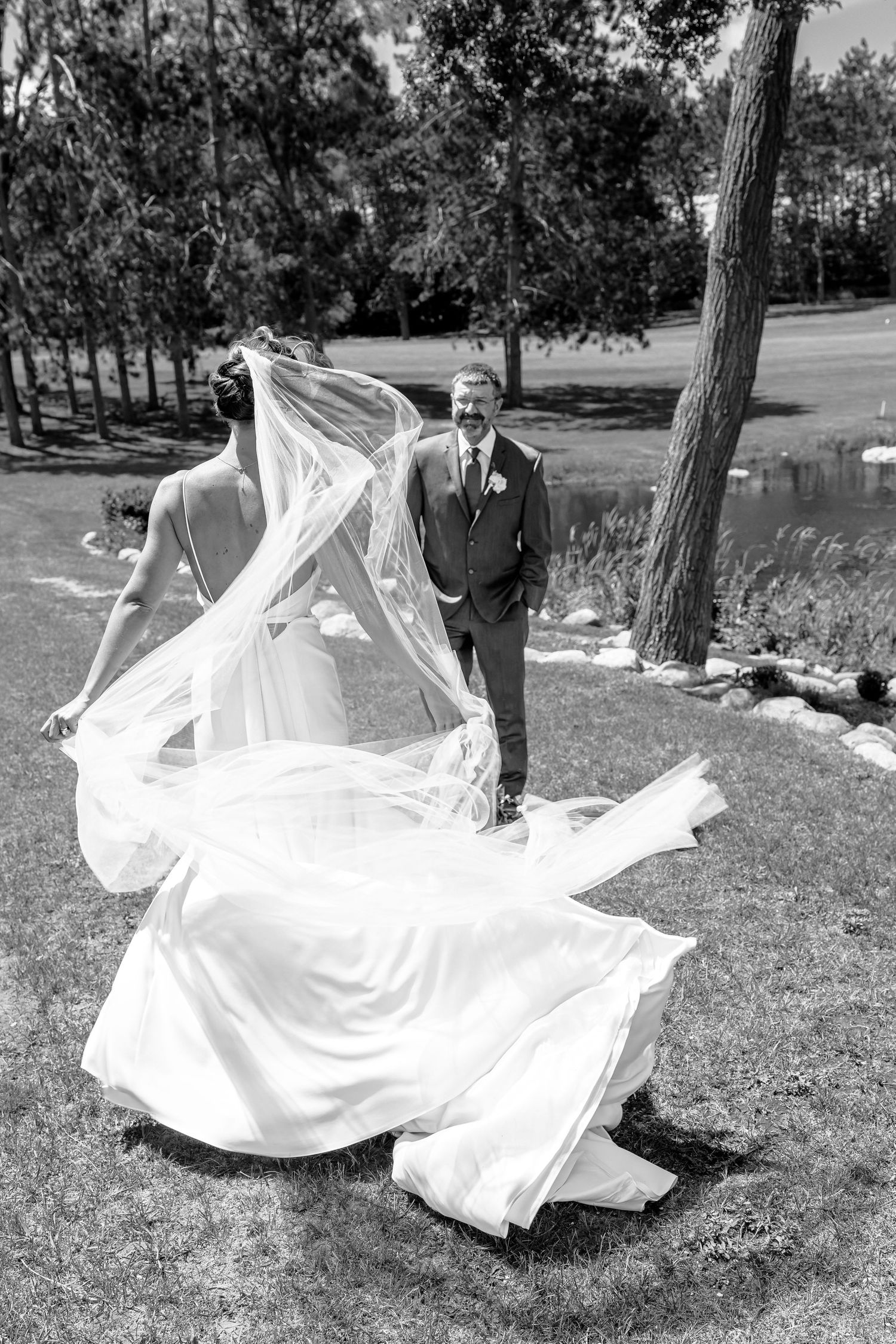 Black and white photo of a bride's veil billowing in the wind as the groom smiles outdoors near trees.
