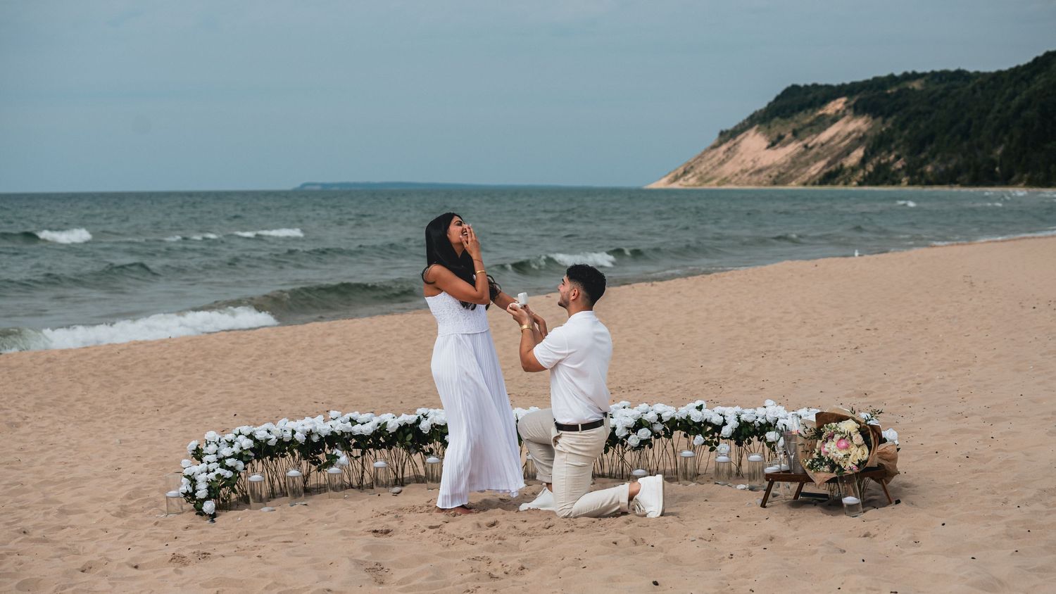 Man proposes to woman on sandy beach with floral arrangement, ocean waves and green cliffs in background.