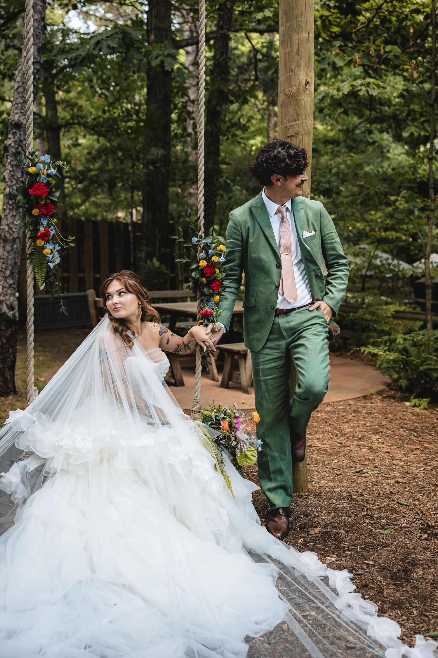Bride in white gown sits on floral swing while groom in green suit stands beside her in a forest wedding setting.