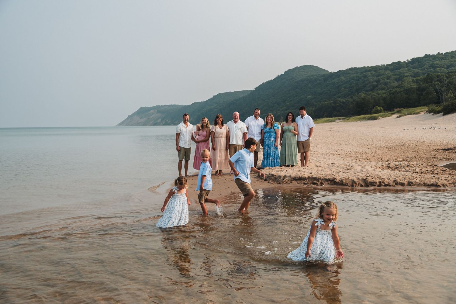 Family group photo on a sandy beach with children playing in shallow water, green hills in the background.