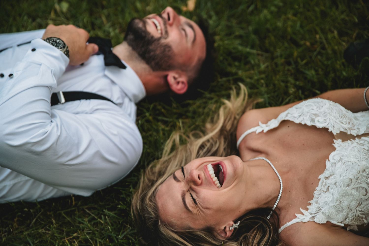 Joyful bride and groom laughing while lying on green grass in wedding attire on their wedding day.