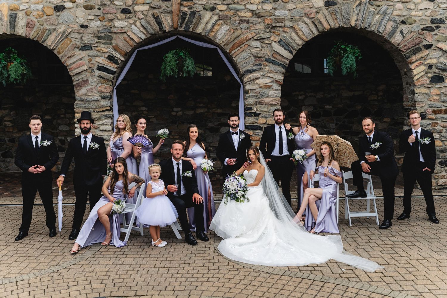 Wedding party poses together on stone steps in front of rustic archway during outdoor celebration.