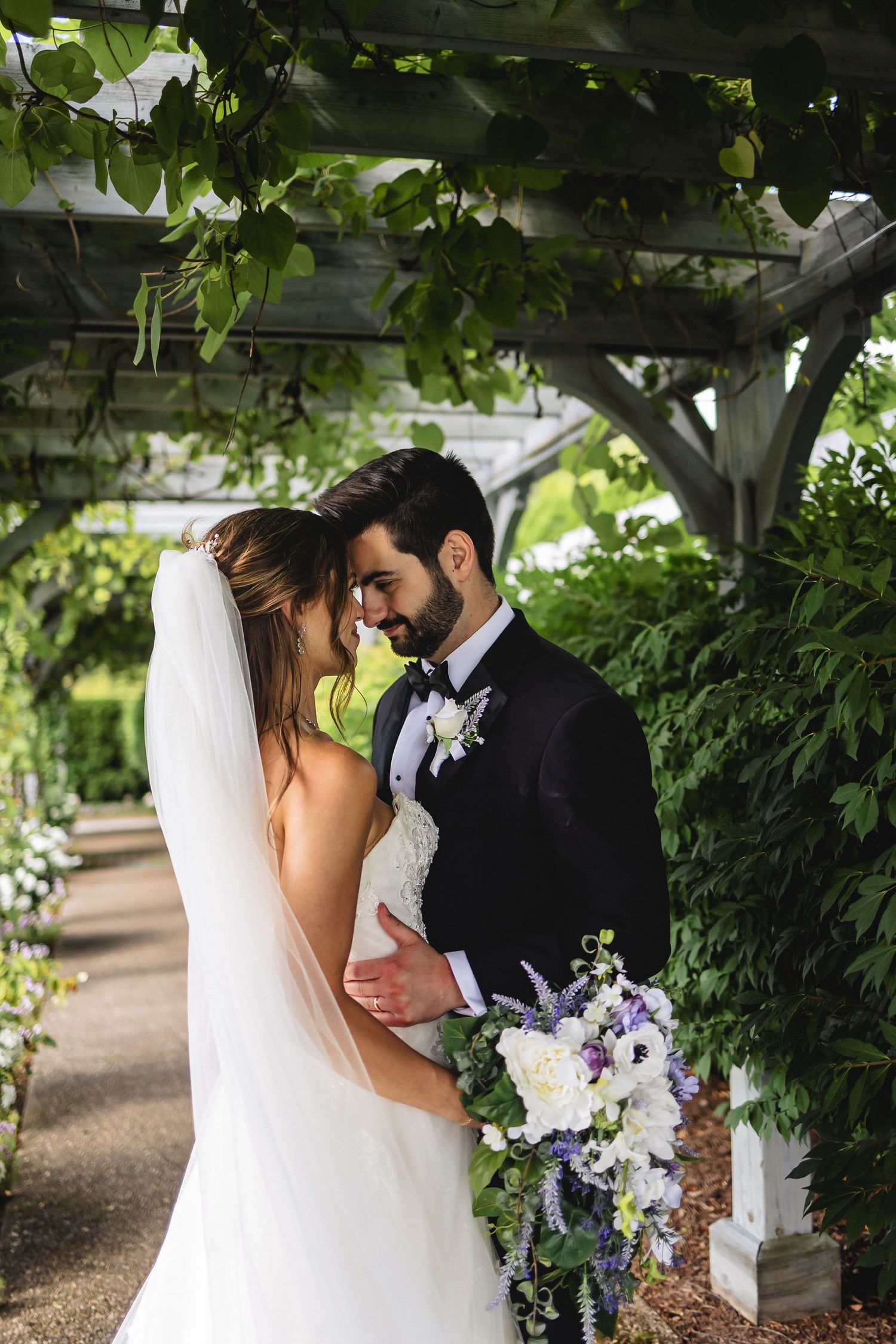 A bride and groom share a romantic moment under a lush garden pergola, with the bride holding a cascading purple and white bouquet.