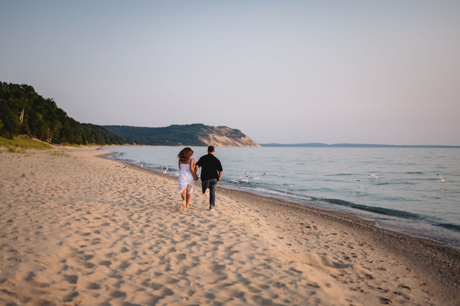 Couple running hand-in-hand along a sandy beach at sunset with calm turquoise water and green hills in the background.