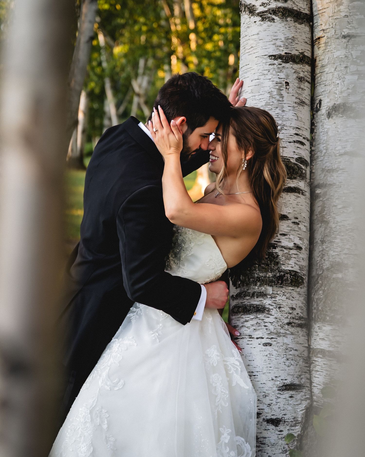 A bride and groom share a romantic moment leaning against birch trees in a lush outdoor wedding setting.