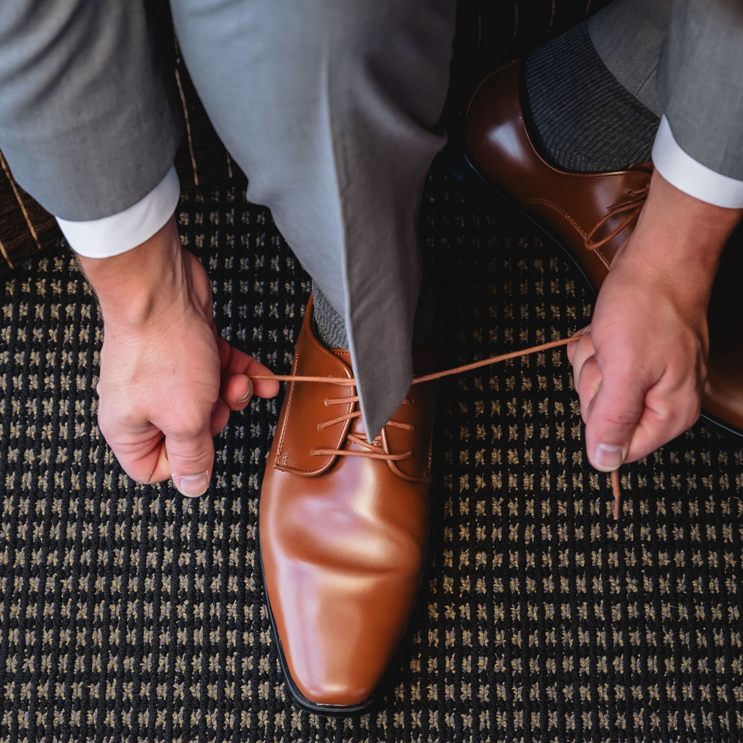 Man tying brown leather Oxford dress shoes while wearing grey suit trousers on patterned carpet floor.