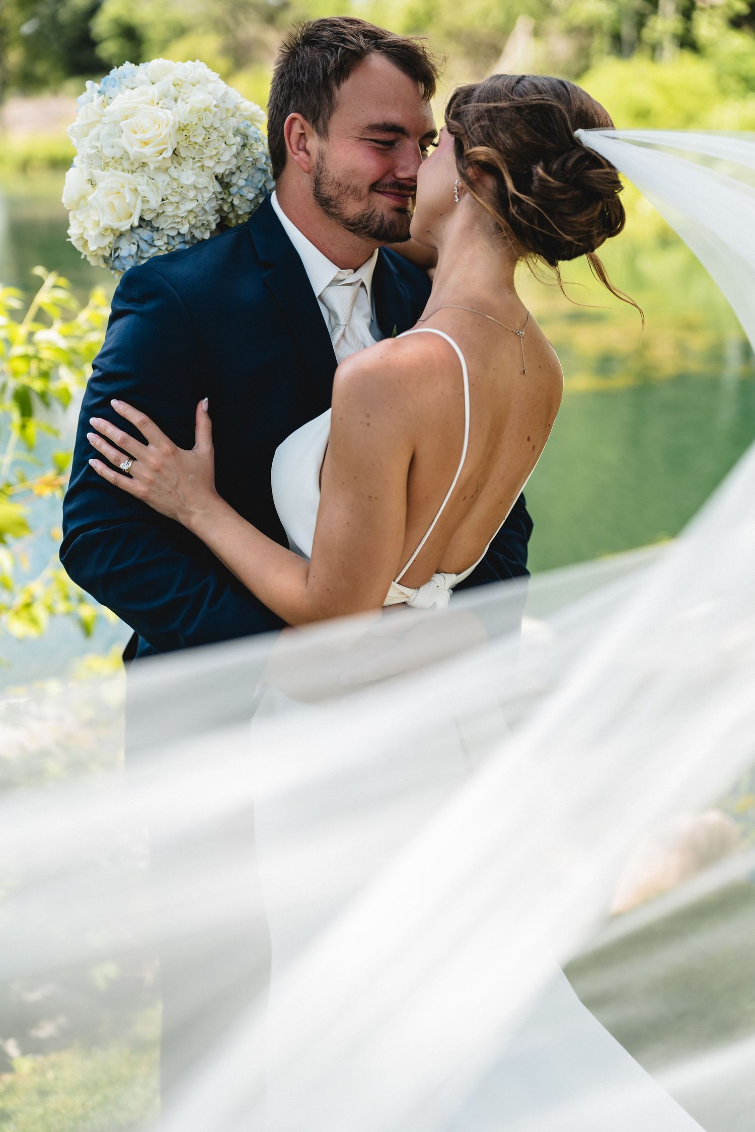 Bride and groom sharing a romantic moment outdoors, her veil flowing, as she holds white hydrangeas in a lush green setting.
