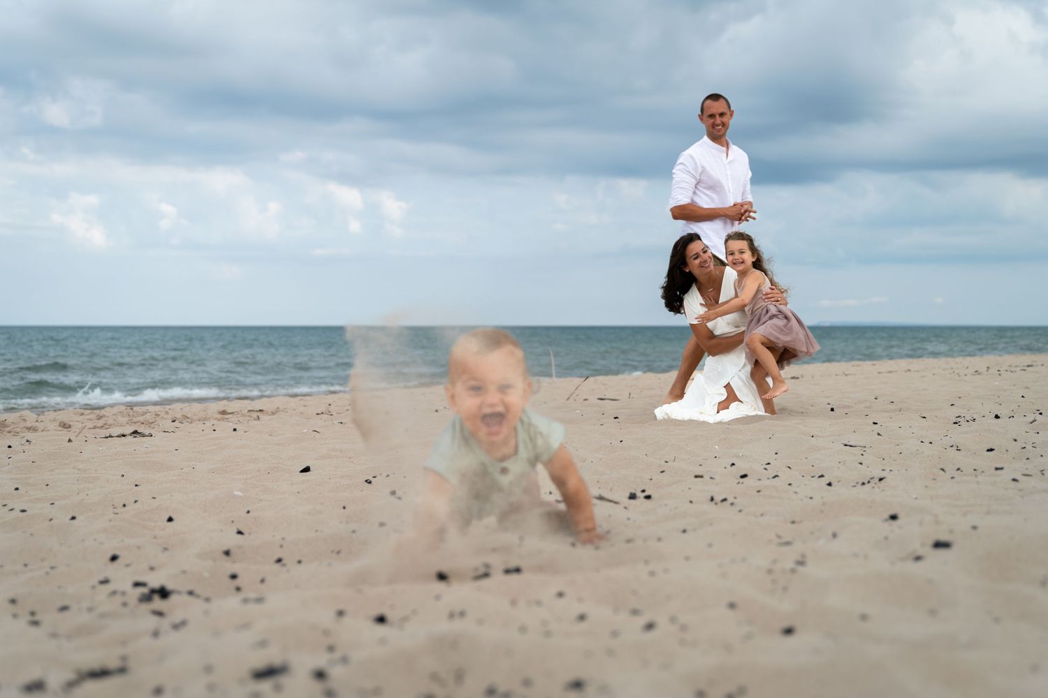 Family enjoying a sunny beach day with baby crawling in sand while parents smile in the background.