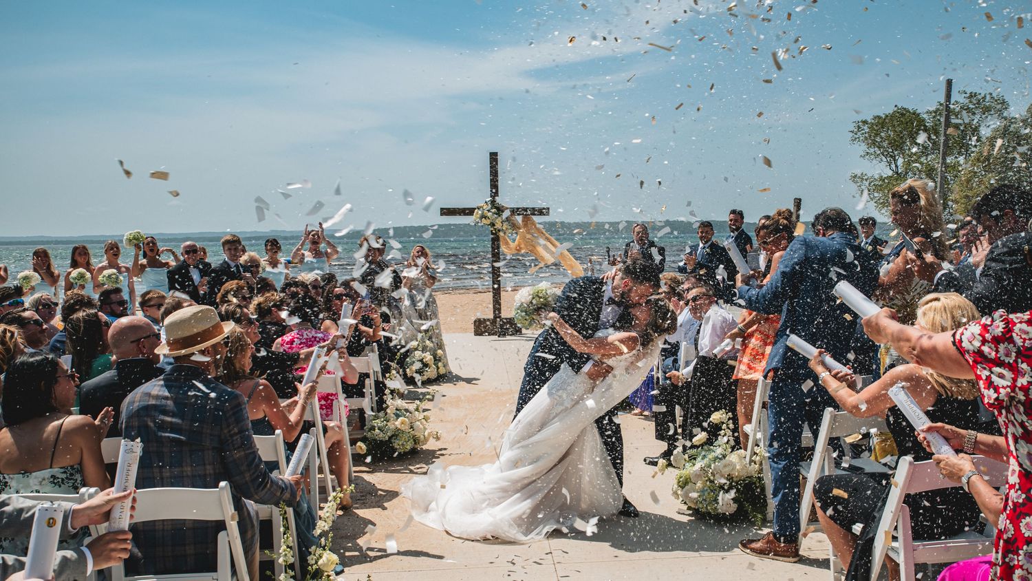 Joyful outdoor beach wedding ceremony with couple kissing as guests toss confetti under bright blue sky.