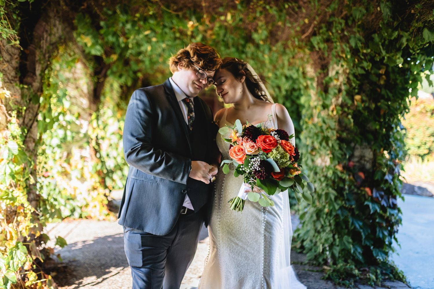 Bride and groom share an intimate moment under ivy-covered arch, bride holding vibrant red and orange bouquet.