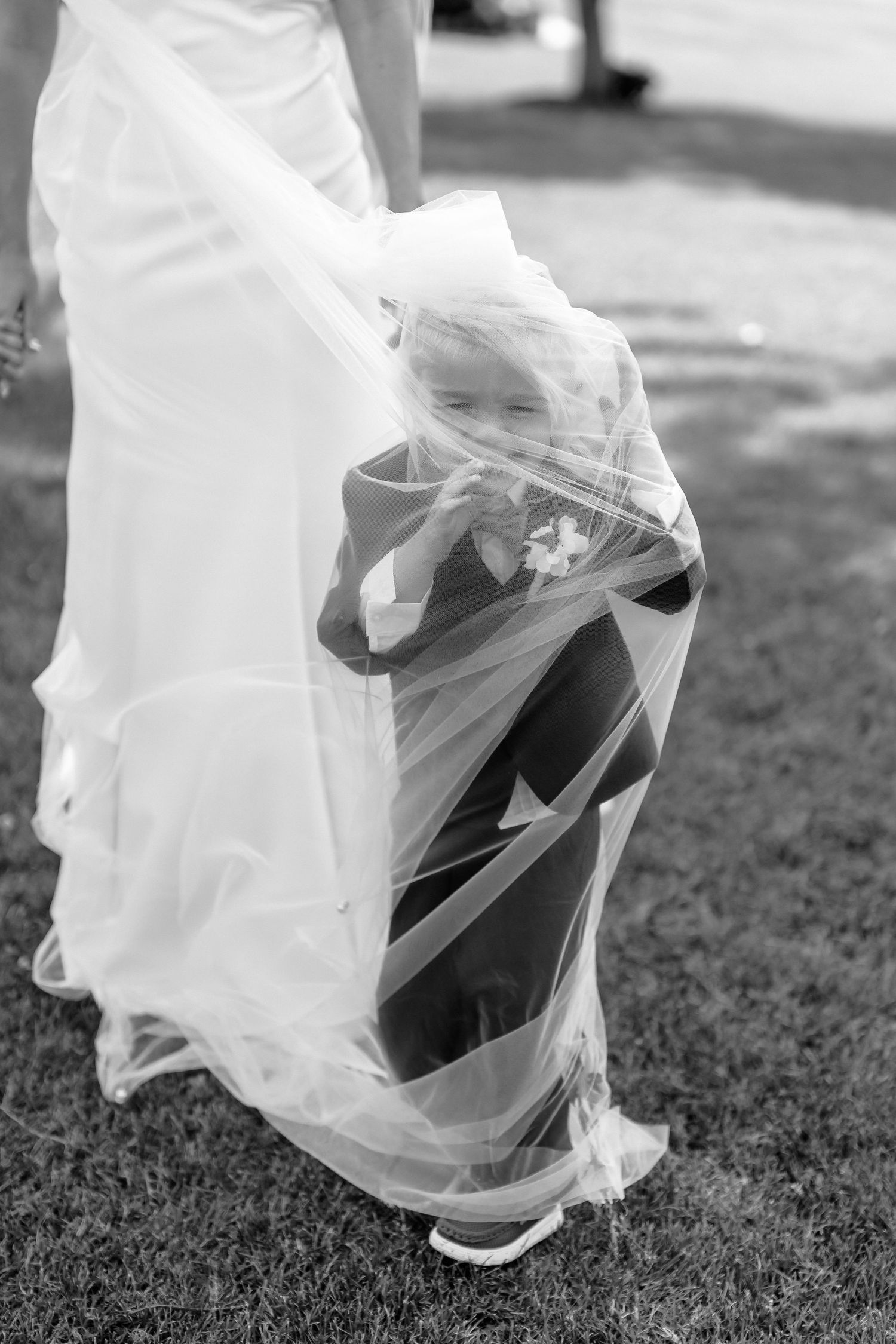 Black and white photo of a child wrapped in a bride's veil at a wedding, smiling while dressed in formal attire.