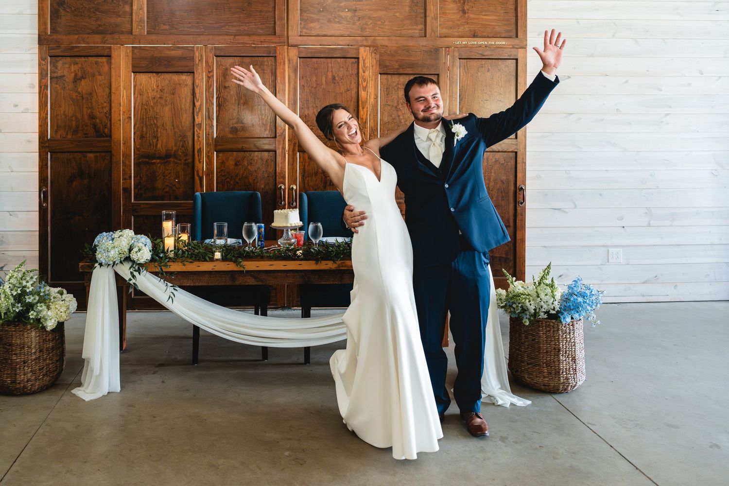 Joyful bride and groom celebrating at wedding reception, arms raised, white dress and navy suit, wooden backdrop.