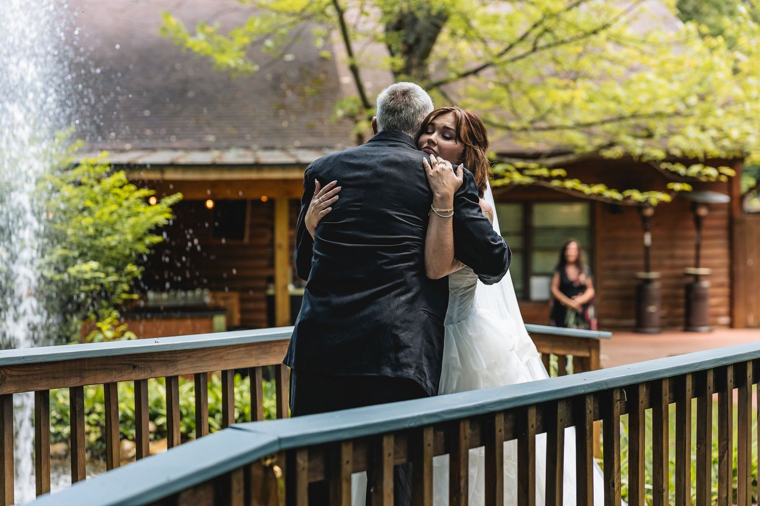 Bride and groom share an emotional embrace on a wooden deck near a fountain, surrounded by lush green trees.