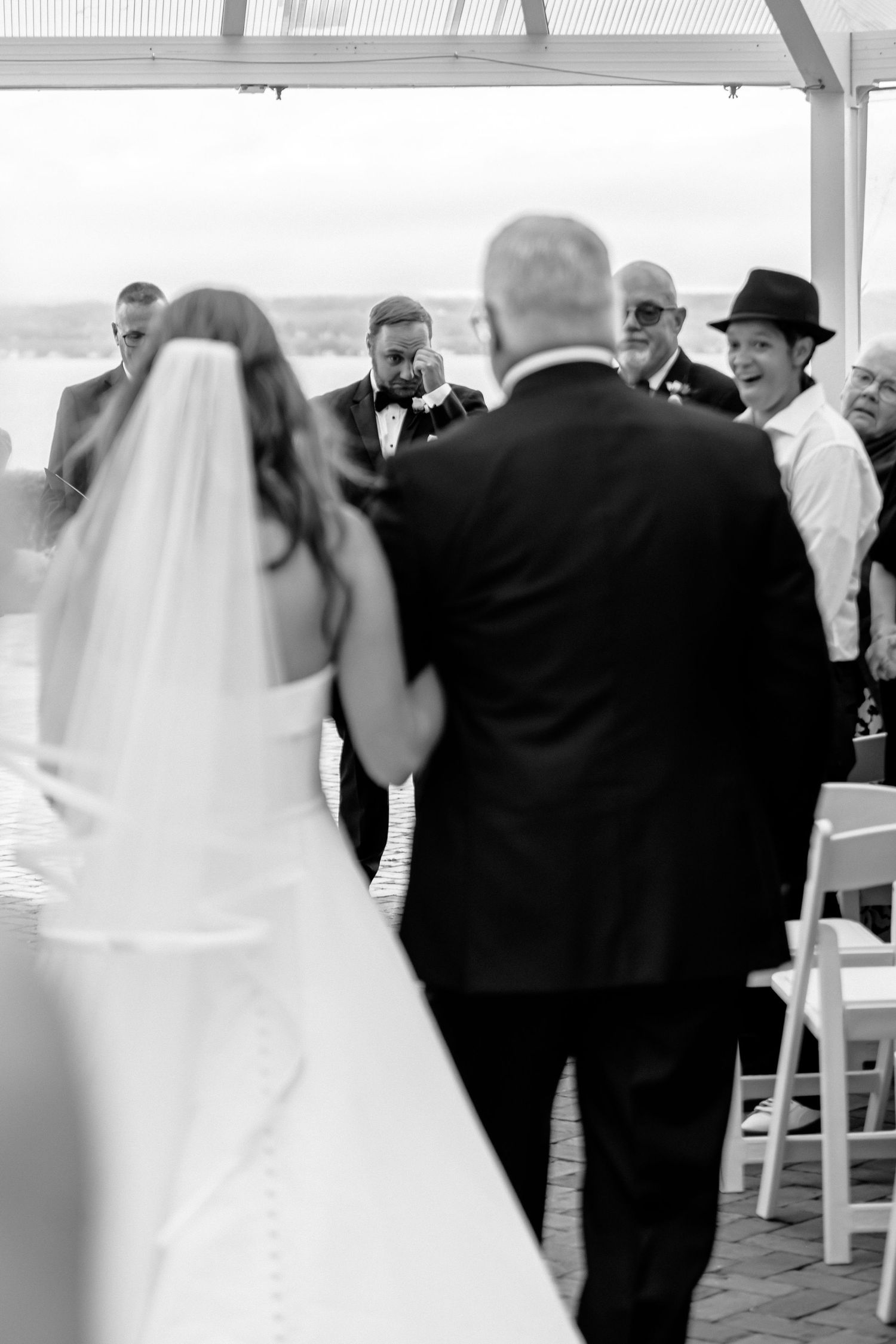 Black and white photo of a bride walking down the aisle with her father toward an emotional groom at an outdoor wedding.