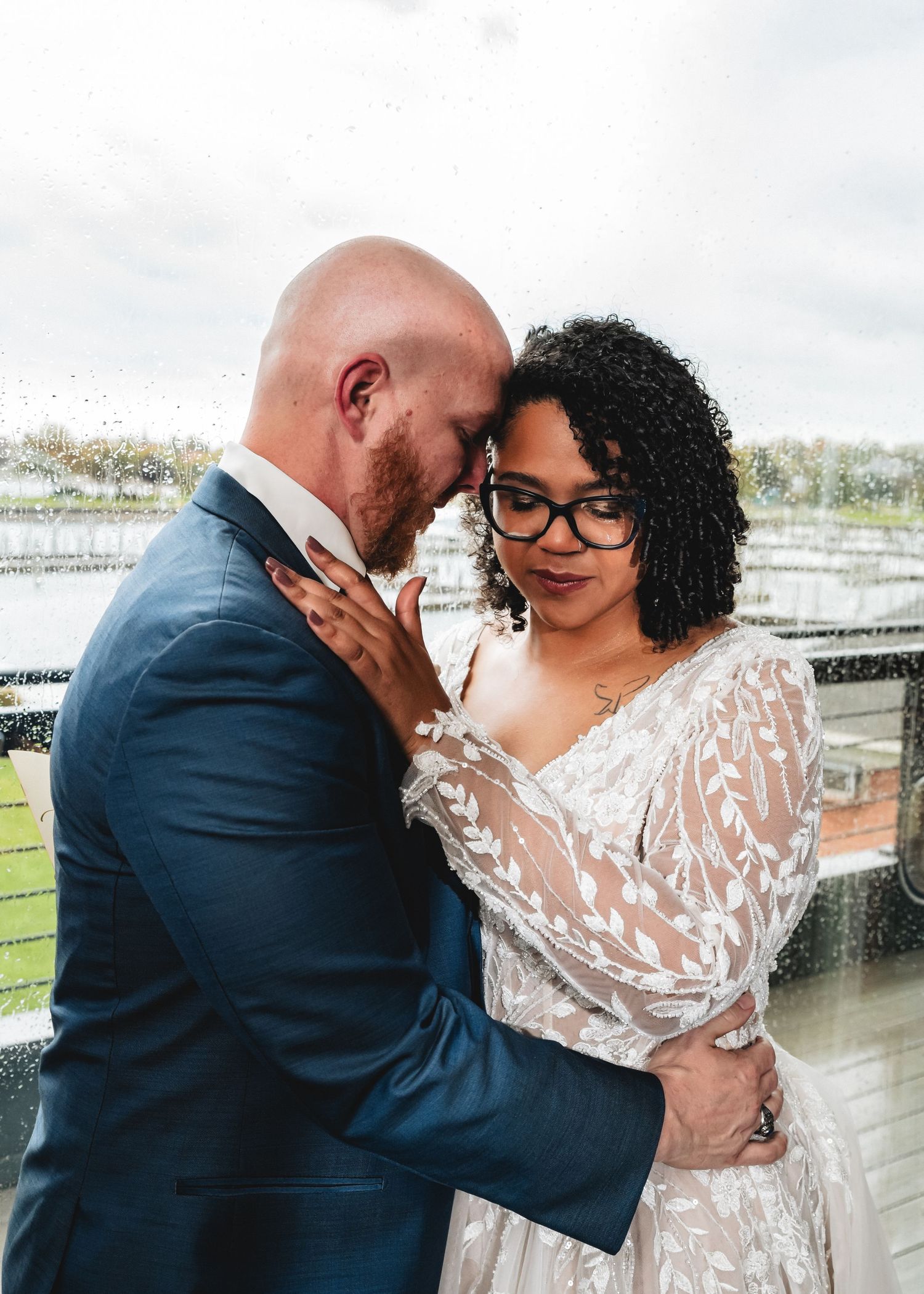 Bride and groom share an intimate moment in the rain, wearing wedding attire on an outdoor venue balcony.