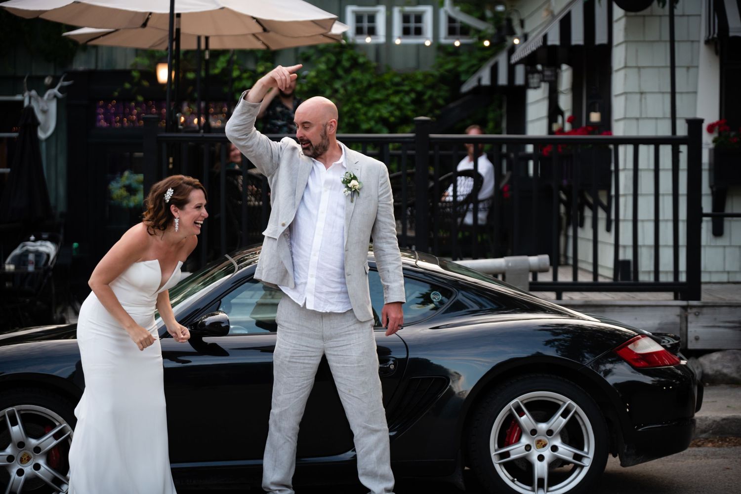 Bride and groom celebrating beside a black Porsche sports car outdoors after their wedding ceremony.