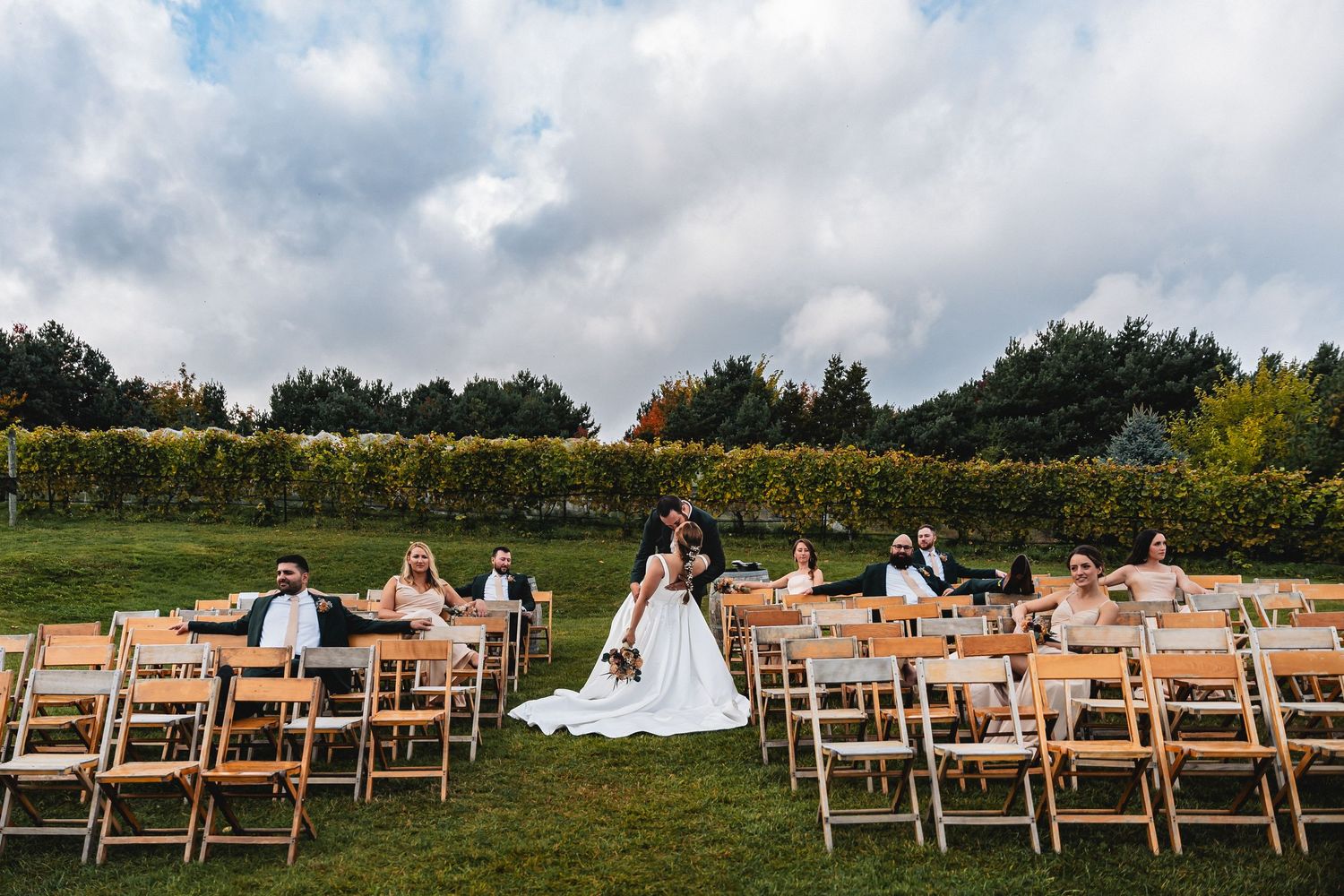 Bride and groom stand at outdoor vineyard wedding ceremony with wooden folding chairs and lush green vines under cloudy sky.