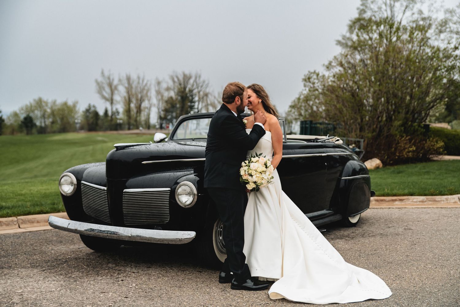 A classic black vintage car serves as an elegant backdrop for a romantic wedding photo shoot on a sunny day.