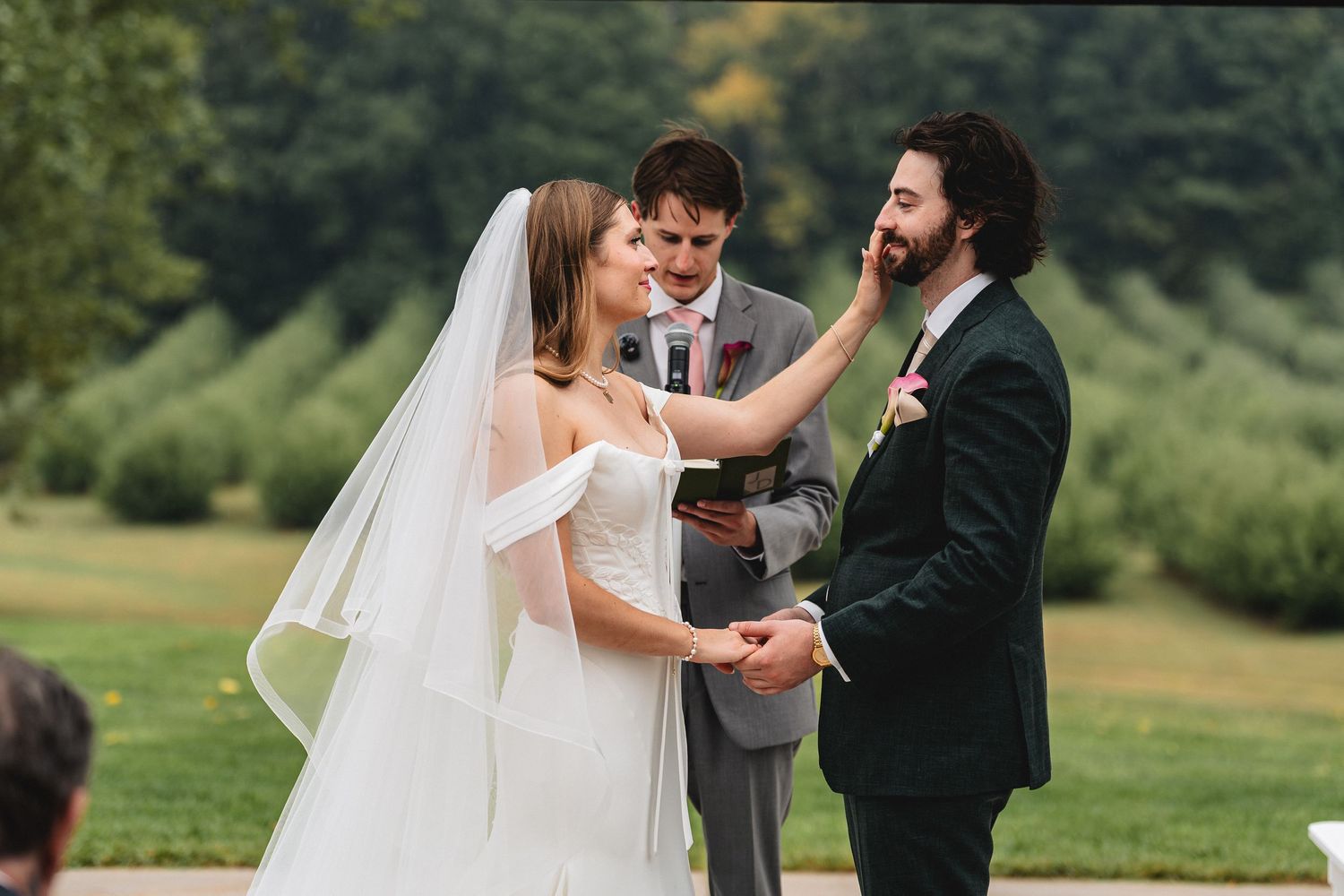 Bride tenderly touches groom's face during outdoor wedding ceremony while officiant holds book in lush green setting.