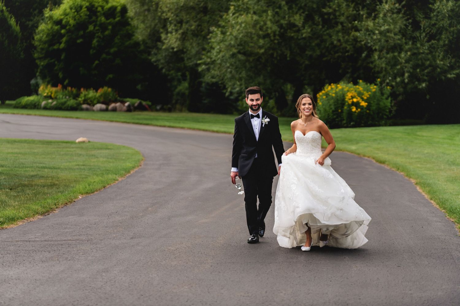 A happy couple walks hand in hand down a winding path on their wedding day, surrounded by lush greenery and gardens.