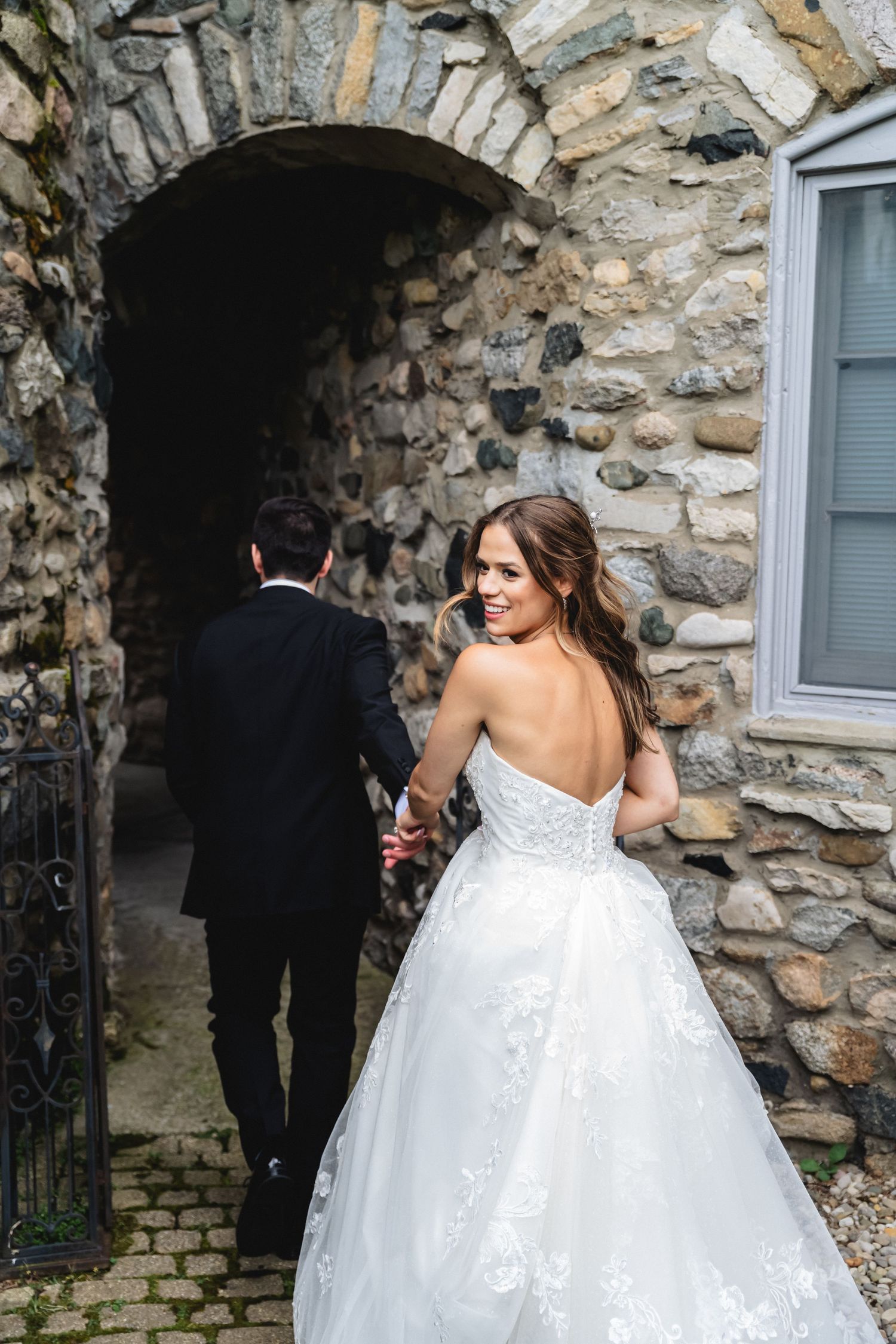 Elegant wedding party walking through a rustic stone archway passage.