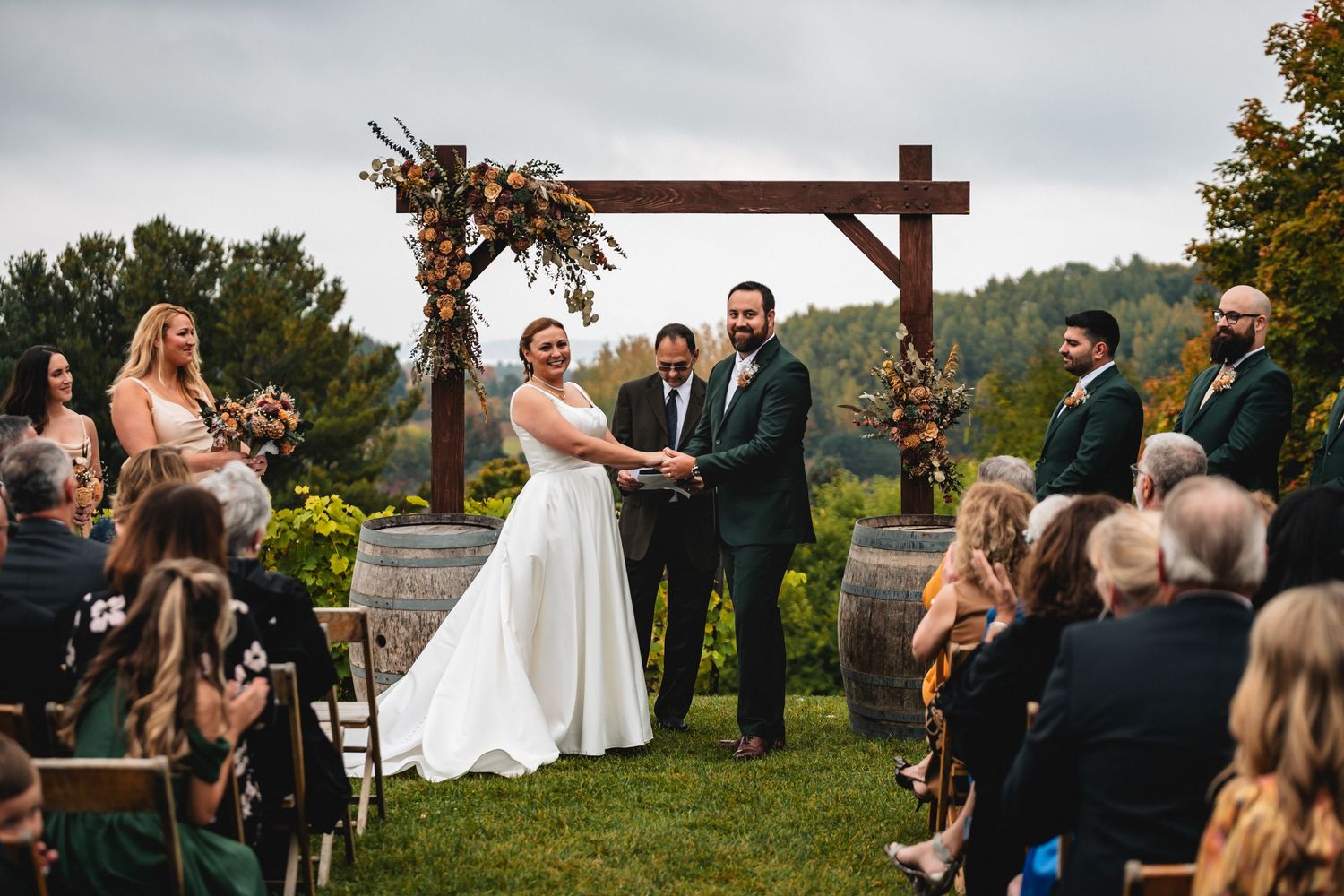 Bride and groom exchange vows at outdoor vineyard wedding ceremony under rustic wooden arch with floral decor.