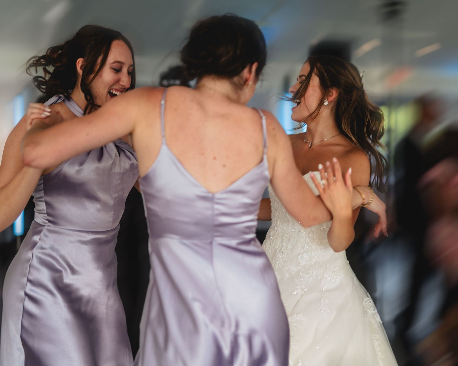 Bride and bridesmaids in lavender satin dresses dancing and laughing together at a wedding reception.