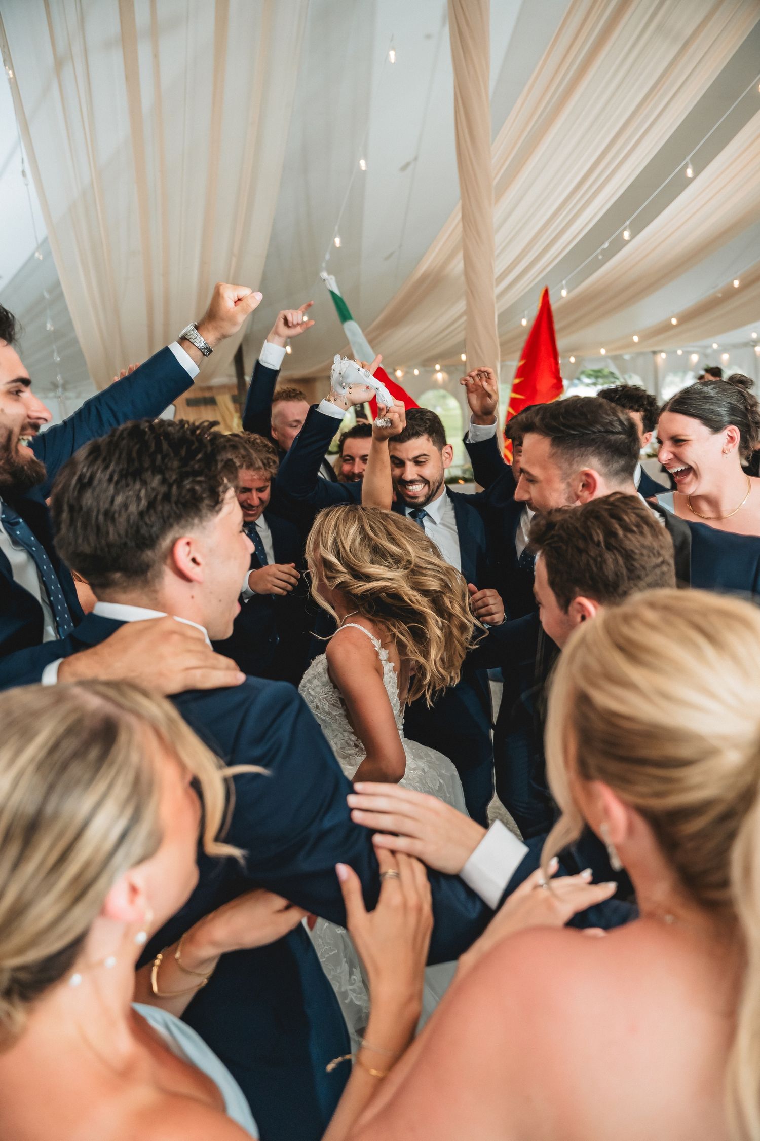 Wedding guests dancing and celebrating on a dance floor under draped white fabric ceiling at a reception party.