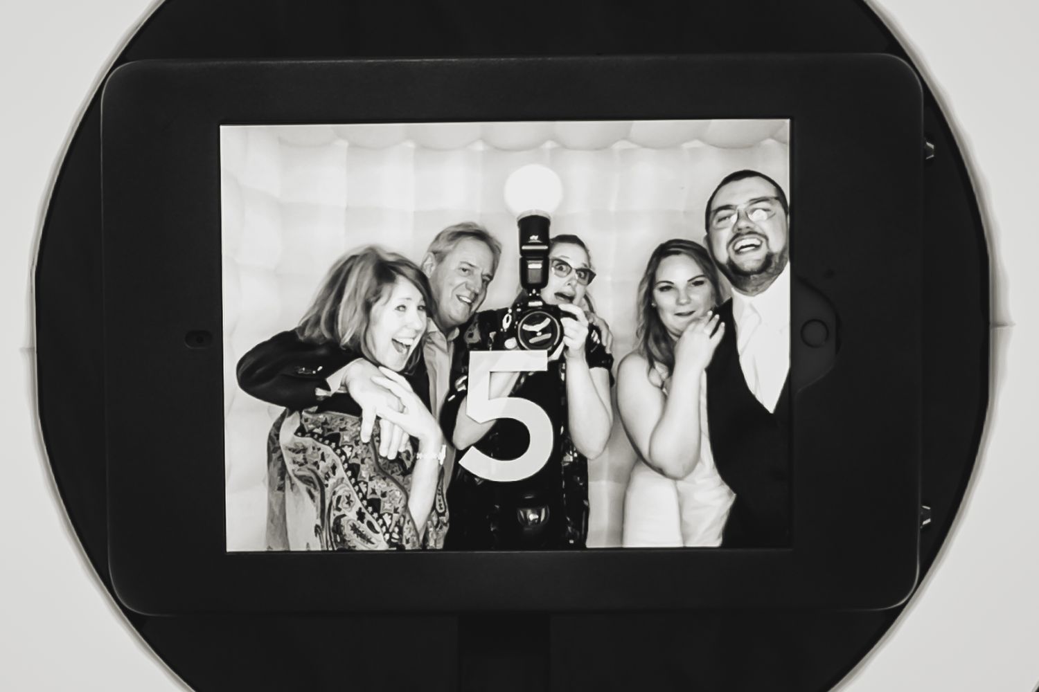 A black and white photo booth strip showing a wedding photographer and newlyweds posing playfully together.