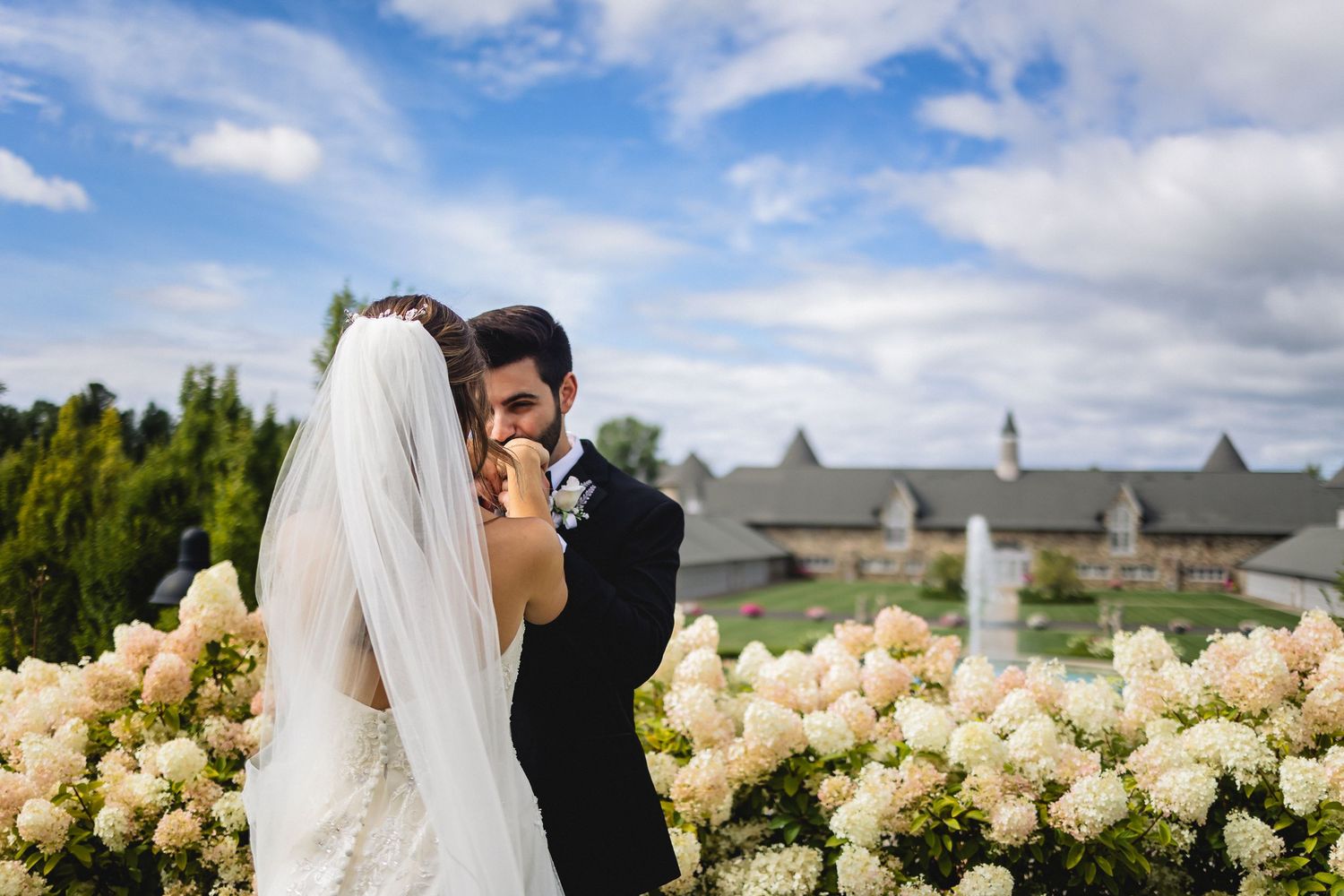 Wedding couple embraces in a garden setting with cream colored roses and a cloudy blue sky background.