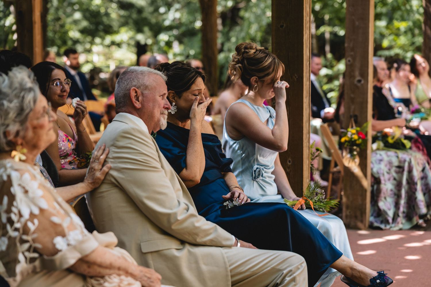 Emotional wedding guests crying and smiling during outdoor ceremony, seated on wooden chairs in a garden venue.