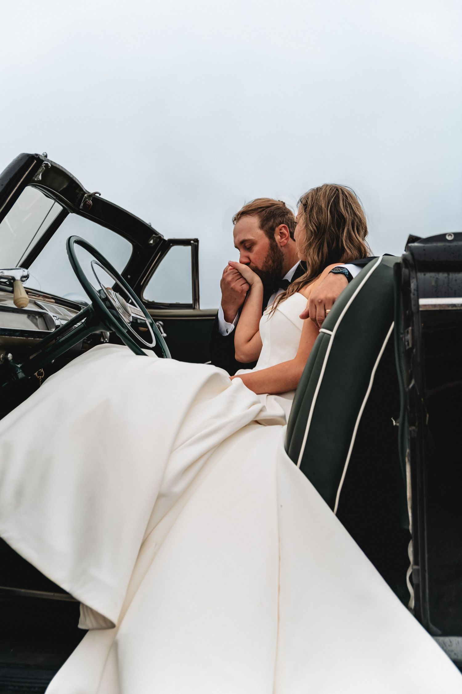 Groom kisses bride's hand as they sit together in a vintage convertible, her long white wedding gown draped over the car at The Hotel Perry