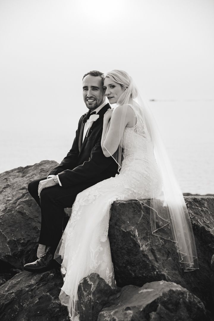 Black and white photo of a smiling bride and groom sitting on rocks by the water in wedding attire.