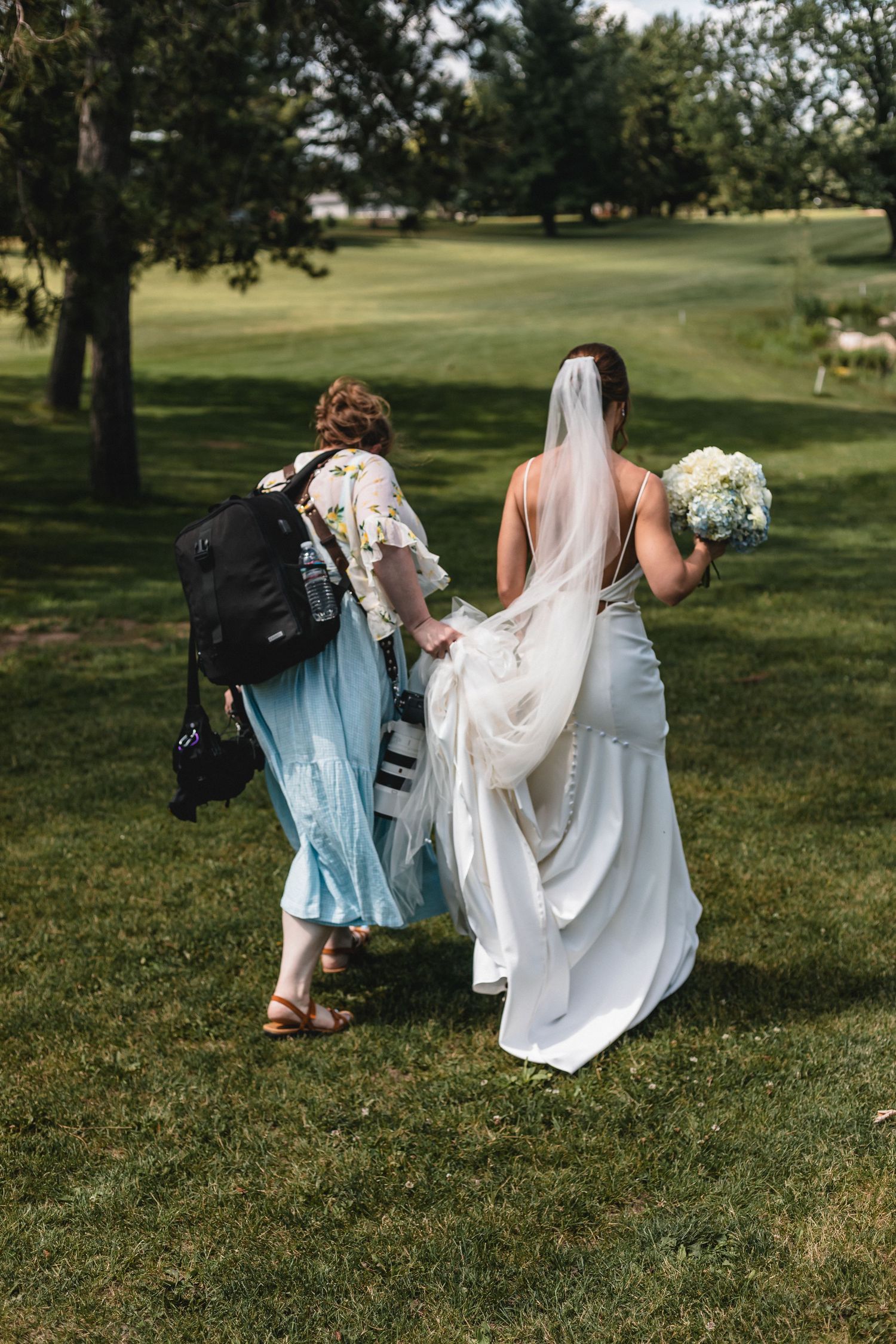 Bride in white gown holding bouquet walks with northern Michigan wedding photographer, Karly Walker, carrying dress train across green lawn.