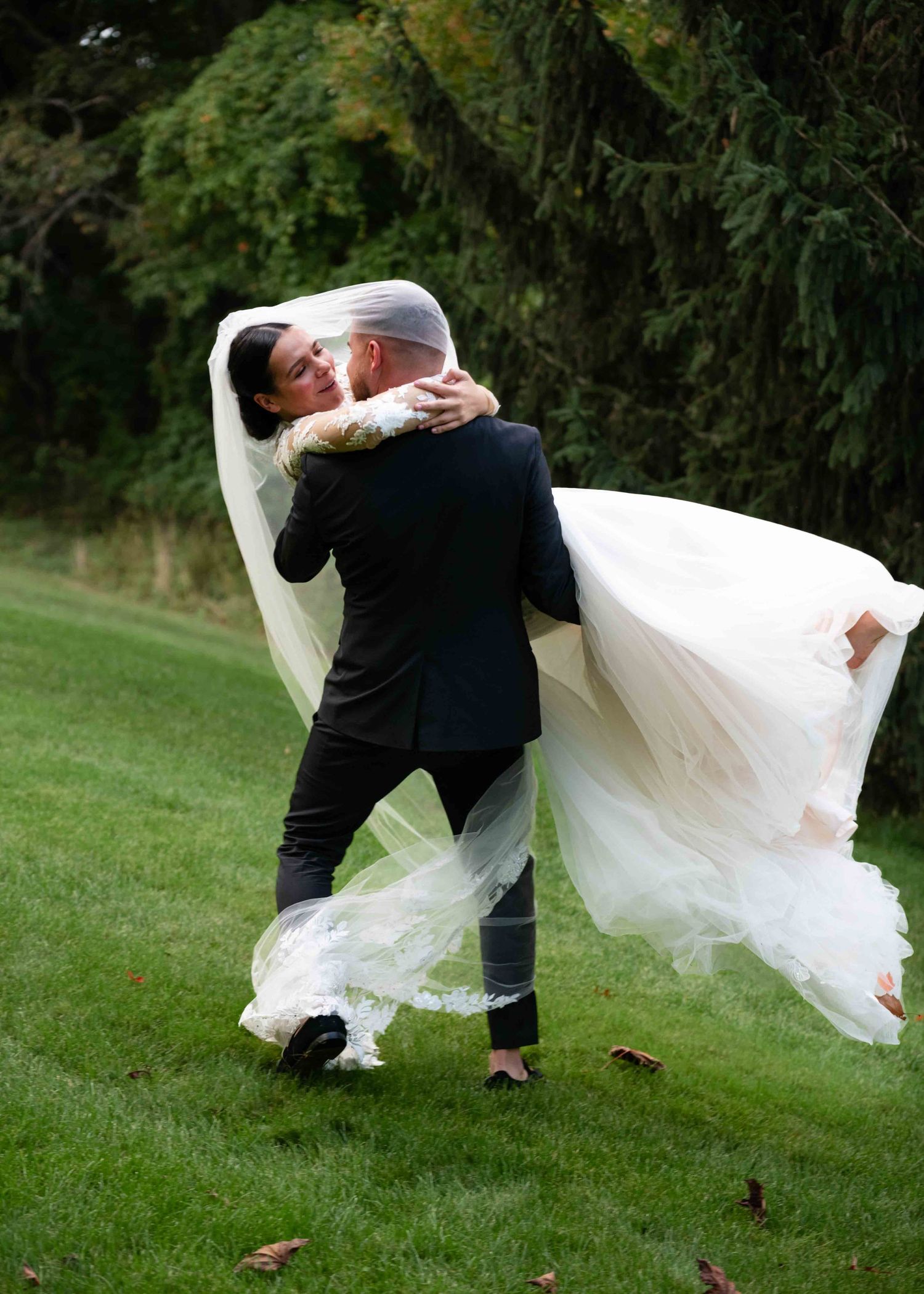 A romantic wedding couple dips and embraces on a lush green lawn surrounded by trees during their outdoor celebration.