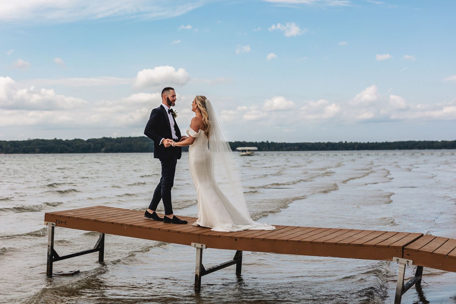 Bride and groom standing on a wooden dock over a lake, holding hands on their wedding day captured by Traverse City wedding photographer
